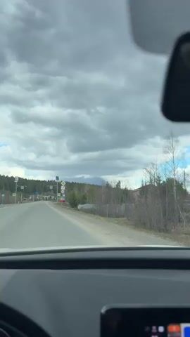 The video is filmed from inside a moving vehicle, looking out through the front windshield onto a two-lane asphalt road. The road has a dashed white line marking the center and appears to be mostly straight.

The sky is heavily overcast with large grey clouds, although patches of blue sky are visible towards the right, suggesting it's daytime.

On the right side of the road, several road signs are visible. Closest to the camera is a yellow diamond sign, indicating a main or priority road. Below it is a circular red-bordered sign with the number "50" in black, signifying a 50 km/h speed limit. Further down the road on the left, multiple square yellow signs with a black pedestrian crossing symbol (a stylized 'A' within a triangle) are visible, some of which are equipped with small solar panels at the top.

The landscape is characterized by dense tree lines and forested areas flanking both sides of the road. The vegetation appears to be a mix of deciduous trees with bare branches and evergreen pines, indicative of early spring or late autumn. Scattered residential buildings, including a distinct brick-colored house with a red roof, can be seen on the right, nestled among the trees. In the distant center, a faint, dark silhouette of a mountain or large hill is visible beneath the clouds.

The immediate foreground shows the interior of the car, including the top of the black dashboard, a portion of the black steering wheel on the left, and part of an infotainment screen on the lower right.

The primary activity is driving forward along the road. No people are visible. The ambient sound consists of road noise. Visible text includes "50" on the speed limit sign.