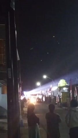 This is a vertical, low-light video of a street scene at night. In the foreground on the left is a dark pole. To the right of the pole are the backs of three people, two facing away from the camera and one facing partially away, looking down the street. In the middle ground, a street is lined with buildings and decorated with what appear to be red and white flags, possibly the Indonesian flag, strung across the street. There are lights illuminating the street, including streetlights and what appear to be stage lights further down. Several vehicles, including what looks like a motorcycle and a car, are visible on the street. In the background, several small, bright, circular lights are visible in the dark sky. The overall atmosphere appears to be festive.
