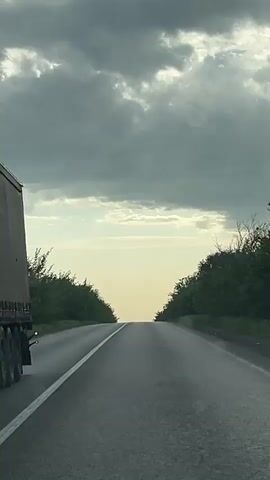 A vehicle drives down a two-lane asphalt road with a single white line dividing the lanes.  Green trees line both sides of the road.  A small black car appears in the distance heading towards the camera.  The sky above is filled with dark gray clouds, with a thin layer of lighter clouds and bright sky visible on the horizon.  The road surface appears wet, possibly from recent rain.