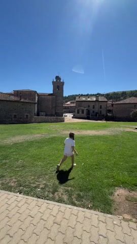 A person wearing a white t-shirt and white shorts is walking across a grassy field on a sunny day. The person is moving away from the camera, towards a cluster of stone buildings. One of the buildings appears to be a church with a tall bell tower. The sky is clear and blue with the sun shining brightly. The grass is green and appears to be well-maintained. The ground in the foreground is paved with interlocking bricks.