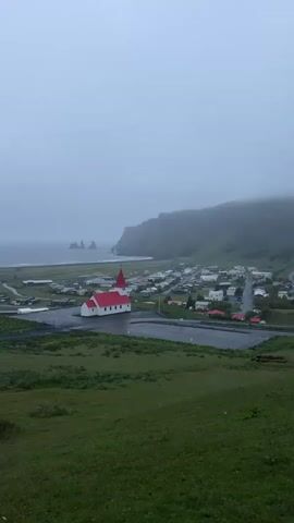 The video shows an overcast landscape featuring a small town by the sea. A red-roofed white church stands prominently in the center of the town. The sea is visible in the distance, with distinctive rock formations emerging from the water. A green, grassy hill slopes downward towards the town. The sky is gray and foggy, obscuring the tops of the cliffs that rise behind the town.