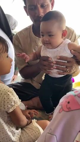 A man is holding a baby, who is wearing a white tank top and black pants. The baby is looking at another child, who is wearing a white top with polka dots. The two children are interacting. The man is wearing a beige shirt. The background appears to be outdoors, possibly under a thatched roof.