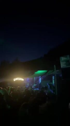 A crowd of people is watching a band perform on a stage at night. The stage is illuminated by bright lights, with a prominent "JAMESON" sign above. The lighting shifts from green to blue and back.  The background features trees and a dark hillside. The camera captures the scene from behind the crowd. People are moving and clapping, suggesting they are enjoying the music. The sky above is a dark blue, indicative of nighttime.  Sound equipment is visible on the right side of the frame.