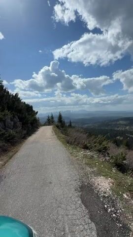 The video shows a paved road leading towards a vista of mountains and sky. The road is grey and appears well-maintained. On either side of the road are low-lying bushes and some small trees. In the distance, a range of mountains is visible, some with patches of snow on their peaks. The sky is blue with many fluffy white clouds. As the camera pans to the right, it reveals a vast landscape of rolling hills and dense forest below the viewpoint.  The landscape includes various shades of green, indicating different types of vegetation. The sun is shining, casting shadows across the scene. There are no people or animals visible.