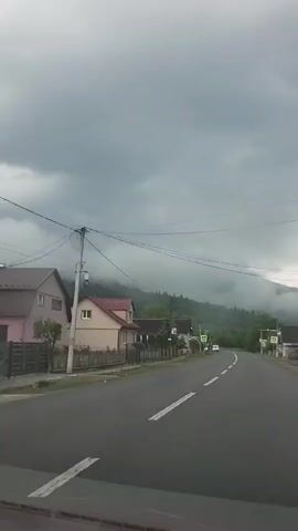 The video shows a road through a village on a cloudy day. The road is asphalt and has white lines marking the lanes. There are houses on either side of the road, and mountains in the background are shrouded in fog. There are two crosswalk signs visible. A truck is parked on the side of the road ahead. The video appears to be taken from inside a moving vehicle.