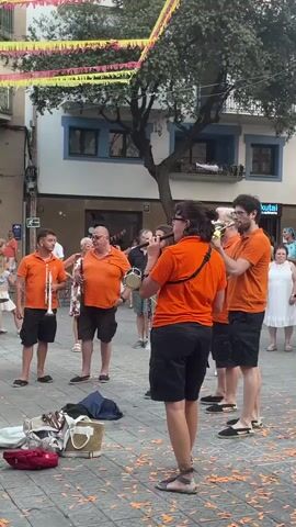 The video captures a street performance with a group of musicians in orange t-shirts playing brass instruments and a percussion instrument. Confetti is being thrown, adding a festive atmosphere. The musicians are positioned in a cobblestone street with buildings and trees in the background, decorated with colorful banners overhead. Several people are observing the performance, some standing and others walking by. There are bags and musical equipment on the ground in the foreground. The focus is on the musicians as they play their instruments.