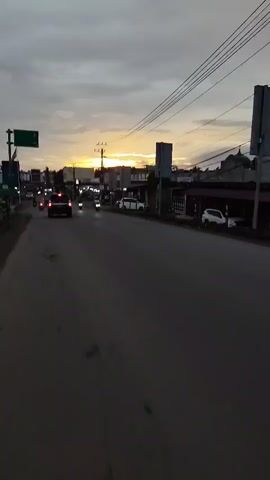 THE VIDEO SHOWS A ROAD IN A TOWN OR CITY AT DUSK. THE SKY IS CLOUDY WITH THE SUN SETTING VISIBLE IN THE HORIZON. THERE ARE SEVERAL VEHICLES ON THE ROAD, INCLUDING CARS AND MOTORCYCLES. BUILDINGS ARE VISIBLE ON BOTH SIDES OF THE ROAD, APPEARING TO BE SHOPS OR HOUSES. THERE ARE ALSO TELEPHONE POLES AND POWER LINES ALONG THE ROAD. THE ROAD ITSELF IS PAVED AND APPEARS TO BE IN DECENT CONDITION, WITH SOME CRACKS VISIBLE. A FEW PEOPLE CAN BE SEEN ON THE SIDEWALK OR NEAR THEIR VEHICLES, BUT THEY ARE NOT CLEARLY VISIBLE. THE OVERALL ATMOSPHERE IS OF A TYPICAL URBAN OR SUBURBAN ENVIRONMENT AS THE DAY ENDS.
