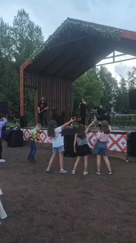 A group of children are dancing in front of a small outdoor stage where a band is performing. The stage is decorated with greenery and red and white patterned fabric. The band consists of at least four members, including a singer and a guitarist. The children are varied in age and attire, with some holding up phones, likely recording or taking pictures. The ground is a dirt or gravel surface, and trees are visible in the background, suggesting a park or outdoor venue. The lighting indicates it's daytime, possibly late afternoon or early evening.