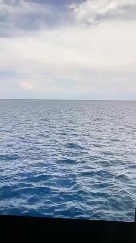 The video captures a view of a large body of water, likely a lake or ocean, with a gentle ripple effect on the surface. The sky above is mostly cloudy, with patches of blue sky visible. In the distance, a range of mountains can be seen, with some vegetation and structures along the shoreline. A few small boats are also visible on the water. The overall impression is one of a serene, natural landscape.