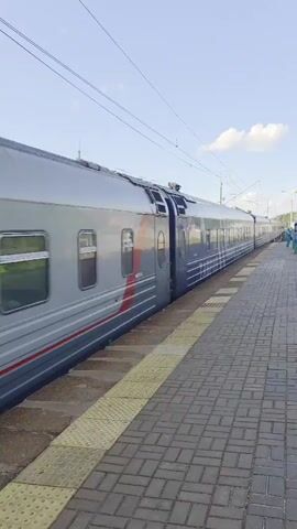 The video shows a train at a station. The train is blue and silver and has the word "РОССИЯ" written on the side. The train is moving. There is a platform next to the train with red and gray paving stones. There are yellow tactile warning strips along the edge of the platform. The sky is blue with some clouds. There are power lines overhead. There is a person in the distance on the platform.