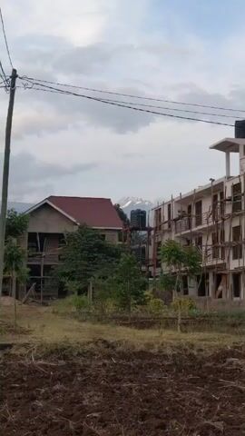 The video shows a landscape with several buildings, trees, and a plowed field in the foreground. In the background, a mountain range with some snow-capped peaks is visible. The sky is overcast with a mix of white and gray clouds.

The buildings are in varying states of construction or completion. One has a red roof and appears mostly finished, while the other building is under construction with wooden scaffolding surrounding it. There are black water tanks on a metal stand situated between the two buildings. 

Trees and vegetation are scattered throughout the scene, adding a green element to the landscape. Power lines are visible stretching across the sky.