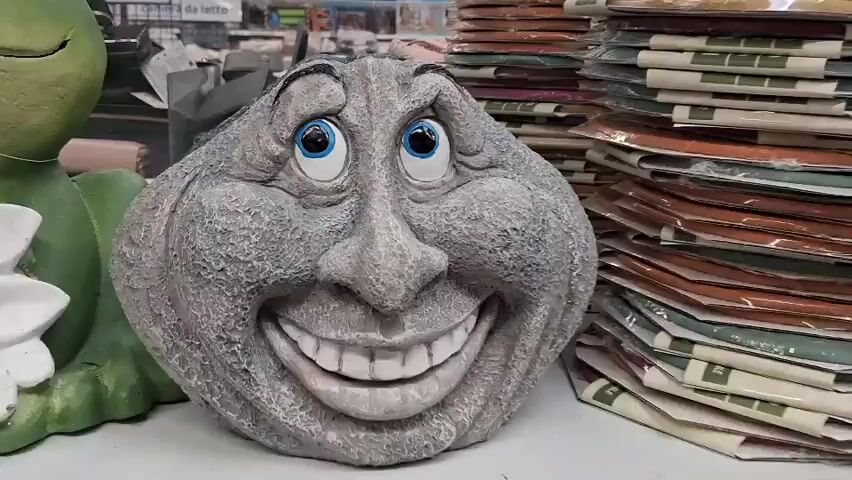 The video shows a close-up of a garden statue of a rock with a carved face. The statue has blue eyes, a large nose, and a wide, toothy grin. The rock is textured to resemble stone and has a slightly rough surface. In the background, there is a green frog statue and stacks of packaged items, possibly craft supplies or home decor, on shelves. The overall scene appears to be a retail environment, likely a store.