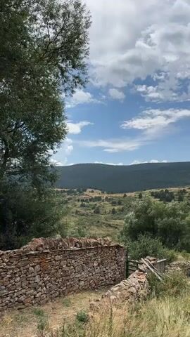 The video shows a panoramic view of a rural landscape under a cloudy sky. In the foreground, a dry stone wall runs diagonally across the lower part of the frame, separating the viewer from a valley. The wall is made of rough-hewn stones of various sizes and colors, and the ground around it is covered in dry grass and some loose stones. To the left of the wall, a large tree with dark green foliage partially obscures the view of the landscape beyond.

The middle ground of the video reveals a sprawling valley dotted with sparse trees and shrubs, with a few small buildings or structures visible in the distance. The terrain appears to be gently rolling, with patches of dry, sun-bleached grass interspersed with areas of greener vegetation.

In the background, a vast expanse of dark green forest covers the hills that form the horizon. The sky is a mix of bright blue and fluffy white clouds, suggesting a partly cloudy day. The overall impression is one of a serene, undeveloped natural environment, possibly in a Mediterranean or similar climate. There are no people or animals visible in the video.