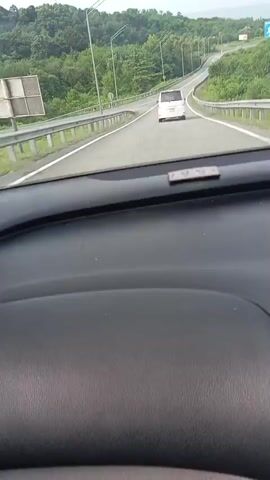 This is a first-person perspective video taken from inside a vehicle. The vehicle is driving on a paved road with white lane markings. On the left side of the road, there is a metal guardrail, and beyond that, a lush green forest covers rolling hills. On the right side of the road, there is also a guardrail, and further in the distance, more trees and a cloudy sky are visible. A white van is driving ahead of the vehicle in the same lane. Another dark-colored vehicle is passing the camera vehicle on the left. The dashboard of the vehicle is visible in the foreground. The video appears to be taken during the daytime under overcast conditions.