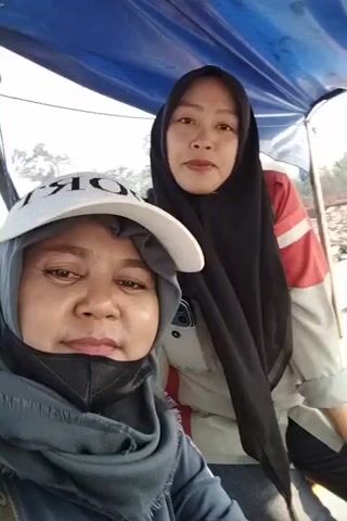 TWO WOMEN, ONE WEARING A WHITE CAP AND HEADSCARF, AND THE OTHER WEARING A BLACK HIJAB, ARE SELFIEING IN WHAT APPEARS TO BE A BOAT. THE WOMAN IN THE CAP IS CLOSER TO THE CAMERA AND IS SMILING. THE WOMAN IN THE HIJAB IS BEHIND HER AND IS ALSO SMILING AND WAVING. THE BACKGROUND SHOWS TREES AND WATER, SUGGESTING AN OUTDOOR, WATERY ENVIRONMENT. THE VIDEO IS SHORT AND SEEMS TO BE A CASUAL MOMENT CAPTURED BY THE WOMEN.