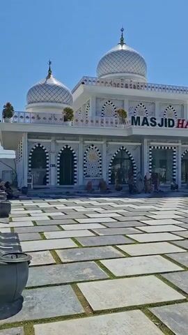 The video pans across the exterior of a mosque on a sunny day. The mosque is white with decorative gold accents and a prominent silver dome. The words "MASJID HARTON" are visible on the building's facade. In front of the mosque is a large paved courtyard with rectangular stone slabs interspersed with grass. Several people are visible in the courtyard and near the mosque entrance, some sitting and some walking. To the right of the mosque, there are trees, benches, and a parked white van. The sky is clear and blue.