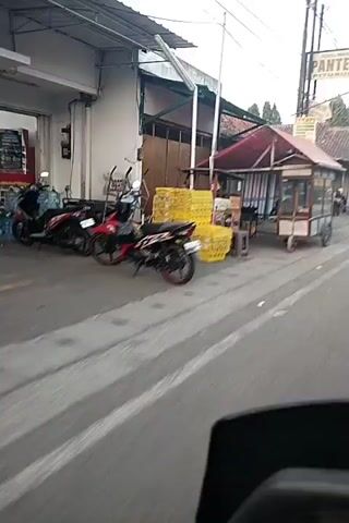 The video captures a street scene, likely in a Southeast Asian country, from the perspective of someone on a moving vehicle. In the foreground, a portion of a dark object, possibly the dashboard or handlebar of the vehicle, is visible. The background features a roadside with several stalls and shops.

At the beginning of the video, a small, mobile food cart with a red awning and a wooden frame is visible. It is parked on the side of the road, and there are stacks of yellow items to its left. As the video progresses, the view shifts to show a row of shops with shuttered fronts, some painted pink and white. A motorcycle is parked in front of one of these shops. Further down the street, a person is seen standing near some boxes.

In the latter part of the video, a person riding a motorcycle passes by, captured in motion blur. The person is wearing an orange shirt and appears to be looking towards the camera. The overall environment suggests a lively street, possibly during the daytime or early evening, with typical urban elements like shops, stalls, and vehicles. The footage is somewhat shaky, indicating movement.