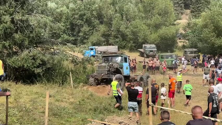 This is a wide shot of an off-road event. In the center of the frame, a large blue and green truck is navigating a muddy incline. The truck appears to be a heavy-duty vehicle, possibly military surplus, with its hood open and some of its front end dislodged. Several people, some wearing high-visibility vests, are gathered around the truck, seemingly assisting or observing. In the background, there are more vehicles, including trucks and vans, and a crowd of spectators. The environment is a grassy, wooded hillside on a sunny day. The spectators are standing behind a rope barrier.
