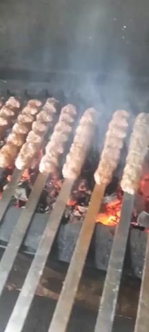 A row of meat kebabs is cooking over hot coals. The kebabs are on metal skewers and appear to be ground meat, formed into elongated shapes. Smoke is rising from the coals and the kebabs, indicating they are actively cooking. The coals are glowing red and orange, providing the heat for grilling. The background is dark and appears to be a grill or fire pit. The video focuses on the food as it cooks.