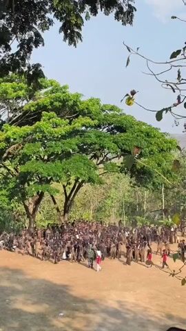 The video shows a large group of people, predominantly males, gathered outdoors in a sandy area with trees and foliage in the background. Many individuals are dressed in similar dark-colored uniforms or clothing. Some are standing in formation, while others appear to be milling about. A few individuals in red and white clothing are interspersed within the crowd. The sky is visible and appears clear. The overall scene suggests a gathering, possibly a training exercise or event.