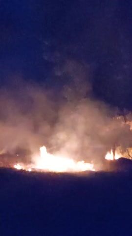 At night, a fire burns in a clearing or field. There are several smaller fires in the vicinity of the larger one. A thin layer of dark grass or dirt is in the foreground. The sky is a dark, dusky blue and a thick layer of smoke rises around the fires. The silhouettes of at least two people can be seen near the fire. They stand motionless. The fire burns brightly, illuminating the area around it.
