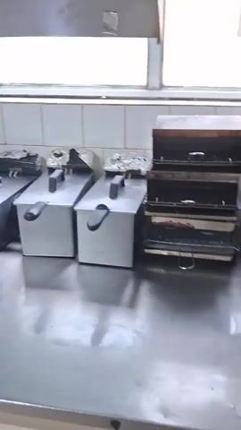 This media file shows a kitchen. The scene is set in what appears to be a commercial or restaurant kitchen with stainless steel countertops and tiled walls. Several pieces of kitchen equipment are visible, including multiple deep fryers lined up, a griddle, and what looks like a gas stove. There is also a sink area with bottles of cleaning supplies and what appear to be utensils hanging on the wall. A blue towel is draped over a countertop. The lighting suggests it is daytime, with natural light coming from a window above the tiled wall. There are no people visible in the footage. The overall impression is of a functional, albeit slightly dated, kitchen space. The location context provided is Marchevo, Bulgaria, which helps to place the scene in Eastern Europe.
