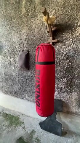 A red boxing bag hangs from a wooden bracket attached to a rough, gray wall. To the left of the bag, a dark, rectangular object is mounted on the wall. The bag has black bands near the top and bottom, and the word "BOXING" is printed vertically in black letters. A black, irregularly shaped object lies on the floor beneath the bag. The floor is concrete and appears damp in places, with patches of green growth. The lighting suggests an indoor or covered outdoor space.