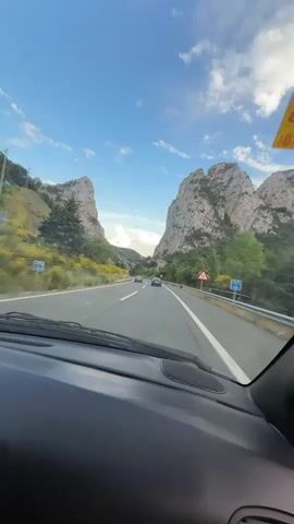 This video is captured from the perspective of a passenger inside a car. The car is driving on a multi-lane highway. On the left side of the highway, there is a steep, rocky mountain slope covered in green vegetation and yellow flowers. On the right side, there is another similar rocky mountain slope with more dense green foliage. The road ahead is a clear asphalt surface with white lane markings. Several cars and a truck can be seen driving in the same direction as the camera's vehicle. The sky is blue with scattered white clouds. There is a triangular road sign with a red border on the right side of the road. The dashboard of the car is visible in the foreground, with a speaker grille on the right side. A yellow sticker with red and black text is attached to the windshield on the top right corner.