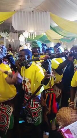 A group of Black men in matching yellow t-shirts, patterned wraparound skirts and hats are playing musical instruments while dancing in a large room. The instruments include long woodwind instruments and drums. The ceiling of the room is decorated with draped white, yellow and green fabrics. People are watching and smiling. The t-shirts feature a design with the words "Family & Friends" and the number "70."