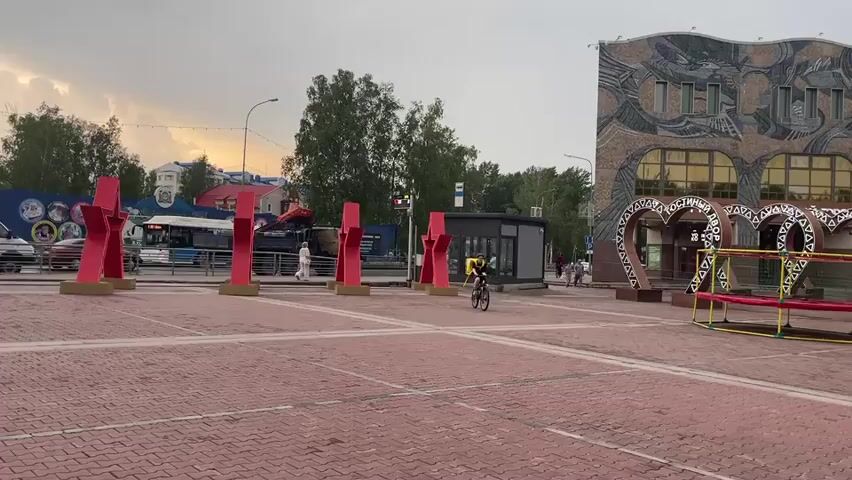 The video shows a wide, open, paved plaza with red brick paving stones. On the right side of the plaza, there is a large, modern building with a mosaic facade depicting abstract shapes and stylized figures. In front of this building, there are three large, decorative heart shapes, and a small playground structure with yellow and red components. A person on a bicycle, wearing a yellow backpack, is riding across the plaza from right to left. To the left of the plaza, there is a street with a bus, a white van, and a military truck. Along the street, there are several tall, red, abstract sculptures resembling stylized figures. Trees and buildings are visible in the background. The sky is overcast with a hint of sunset colors.