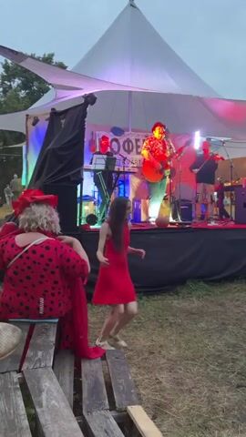 At dusk, on a grassy field under a large white tent, a band performs on a raised stage. The tent is illuminated with colorful lights, casting green, pink, and red hues. The band consists of at least four musicians. One person in the center plays an acoustic guitar and sings, wearing a patterned shirt and a hat. To their left, someone plays a keyboard. To the right, another person appears to be playing drums, and further to the right, another musician stands with an instrument. In the foreground, two women are visible. The woman on the left wears a red dress with black polka dots and a large red hat with a bow. She is facing away from the camera, towards the stage, with a red scarf draped over her shoulder. The woman on the right, closer to the stage, wears a short red dress and is dancing. She has long dark hair and is facing the stage, with her arms outstretched. In front of them, wooden benches are visible, suggesting an audience seating area. The overall atmosphere appears to be a casual outdoor concert or festival.