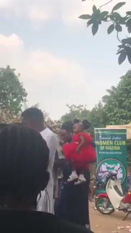 Here's a description of the video:

The video shows a man speaking into a microphone, addressing a crowd outdoors. He's wearing a white outfit. The crowd consists of adults and children. A woman is holding a young girl in a red dress, who also appears to be part of the audience. There's a banner in the background with writing on it, along with a small bicycle. The environment appears to be a yard or garden area with trees. The camera moves around, showing the speaker from different angles and capturing the reactions of the crowd.