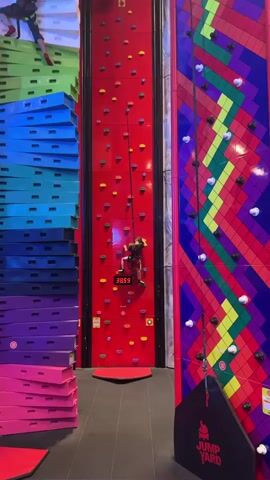 A young boy, secured by a safety harness and rope, is climbing a red rock-climbing wall. The wall has colorful climbing holds and a digital timer displays his climbing time. Beside the red wall is a stack of padded mats of varying colors: green, blue, purple, and pink. To the right of the red wall is a colorful climbing wall with a geometric pattern. The walls are located in a place called “Jump Yard,” as indicated by the sign at the bottom of the geometric-patterned wall. Other people can be heard in the background.