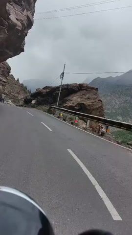 The video shows a POV shot from a motorcycle on a winding mountain road. The road is asphalt with a white line down the center. The motorcycle is moving downhill. On the left side of the road, there is a steep, rocky cliff face. On the right side, there is a drop-off with a metal guardrail. Two trucks are driving ahead on the road. The sky is cloudy and overcast, and mountains can be seen in the distance.