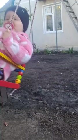 A young child sits in a colorful swing in a backyard. The child wears a pink and white checkered jacket, patterned leggings, a black beanie, and white shoes, and has a pacifier in their mouth. The swing hangs from ropes attached to a metal frame. Behind the swing is a light-colored house with a window and a metal staircase leading to the roof. There is also a tree with white blossoms visible on the left. The ground is dark and patchy, appearing to be dirt or muddy soil. The child is swinging gently back and forth.