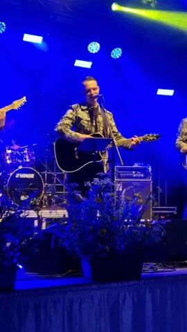 A live music performance is captured in this video. A man in a camouflage jacket is singing and playing an acoustic guitar on a stage illuminated by bright blue lights. He is positioned in the center of the frame, with a music stand in front of him. To his left, a drummer is visible playing a drum kit with "SONOR" branding. Another guitarist, partially visible on the far left, plays an electric guitar. Behind the main performer, amplifiers and other stage equipment are present. In the foreground, a potted plant with dark foliage and small flowers adds a natural element to the scene. The overall atmosphere appears to be energetic and engaging, as suggested by the lighting and the performer's actions.