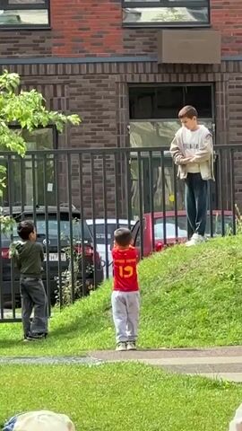 Three young boys are playing outdoors in a grassy area in front of a brick building. A fourth child, partially visible in the foreground, is also present. The boy in the center, wearing a red jersey with the number 19, is facing away from the camera. To his left, another boy in a green shirt is facing the same direction. A third boy, standing on a grassy incline to the right and slightly behind the other two, is wearing a beige jacket and jeans. He appears to be interacting with the other boys, possibly throwing something or gesturing. A black car and a red car are visible behind a black metal fence. Trees and foliage are also present in the scene.