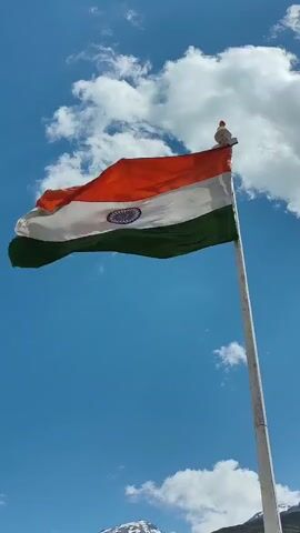 A large Indian flag is displayed on a flagpole against a backdrop of a blue sky with white clouds. The flag, which is made of three horizontal bands of saffron, white, and green, with a navy blue Ashoka Chakra in the center of the white band, is billowing in the wind. Below the flag, a mountainous landscape can be seen, with green vegetation and rocky terrain. The sun is shining, and the scene appears to be set outdoors in a natural environment. The video is shot from a low angle, looking up at the flag.