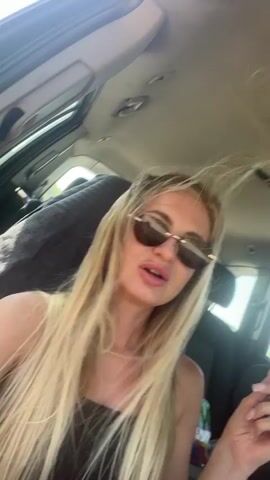 The video shows a blonde woman in sunglasses sitting in the passenger seat of a car. She is holding a small electronic device, possibly a vape pen, in her right hand. The car's interior is visible, including the ceiling and a sun roof. The woman is speaking, and her lips are pursed as if she is talking or smoking. The background outside the car window is blurred, suggesting motion.