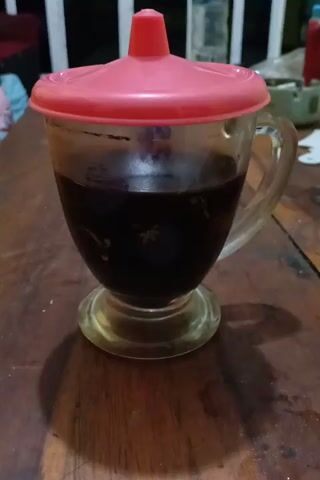 A cup of dark liquid sits on a wooden surface. The cup is made of clear glass with a handle. It has a pink lid with a red spout. The liquid is dark brown and appears to be coffee. The wooden surface is a table with visible grain and texture. The lighting suggests an indoor setting, possibly with a window to the left casting a shadow of the cup onto the table.