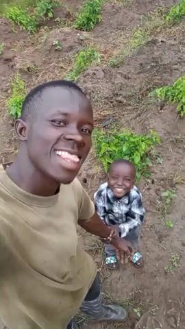 A man and a young child are taking a selfie in a garden. The man is in the foreground, smiling and sticking out his tongue. The child is in the background, also smiling and sticking out their tongue. They are both wearing casual clothing. The garden appears to be on a hillside, with rows of plants in the background. The ground is dry and dusty.