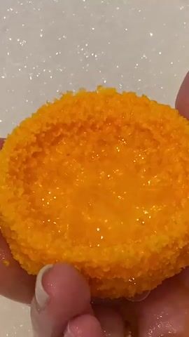 In this close-up video, a round, bright orange object resembling a bath bomb is held by two fingers. The object is textured with small, granular beads, and a clear, viscous liquid fills a concave section in its center. The background is a blurred expanse of white, speckled with tiny, shimmering highlights, suggesting a foamy or bubbly surface, possibly water. The lighting appears bright, creating reflections on the liquid within the object, and a general sense of cleanliness and perhaps relaxation. The skin of the fingers holding the object appears clean and manicured. There is no visible text in the video. The context of "Новое Шигалеево, Russia" places this scene in a residential or personal care setting within that geographical location, likely a bathroom.