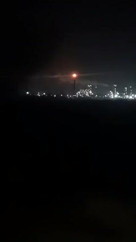 The video shows a distant industrial complex at night. The scene is very dark, and the foreground is completely black. The complex consists of several structures and pipes illuminated by white lights. There is a taller tower with a red light atop it, and some smoke or steam is visible above the complex. The conditions appear hazy, obscuring some details.