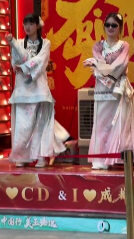 Two young women are performing a coordinated dance routine on a small, elevated stage. They wear traditional Chinese garments, one in pastel blue and the other in pastel pink. The dancer on the right wears sunglasses. The backdrop behind them is a large red banner with gold Chinese characters. An air cooler is placed behind them. The stage is decorated with red and gold hearts and the words "I ❤️ CD & I ❤️ 成都." A metal stanchion with a red ribbon serves as a barrier in front of the stage. The lighting is bright and the overall setting suggests a promotional event or street performance.