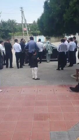 A group of men in white shirts and black pants, along with a few individuals in black clothing, are gathered around a black car. One man in a blue shirt is standing near the car door, appearing to interact with someone inside. A young boy in a black shirt and white pants stands in the foreground, facing away from the camera. The scene takes place outdoors on a paved area with a tiled border, under a bright sky with scattered clouds and trees in the background. A utility pole is visible to the left.