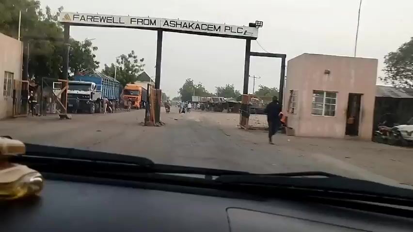Here is a description of the video:

The video appears to have been taken from inside a vehicle driving through the entrance gate of "ASHAKACEM PLC."  The camera looks through the windshield as the car passes under a large sign that reads, "FAREWELL FROM ASHAKACEM PLC".  Beyond the gate, the road is unpaved and dusty. Several trucks are parked along the side of the road. People are standing and walking around the trucks. Some small buildings and structures can be seen in the distance. The overall scene suggests an industrial or commercial area on a somewhat overcast day.