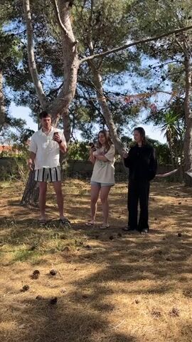 Here's a description of the video:

Three young people are standing in a yard or grassy area with pine trees. The ground is covered in pine needles and pine cones. The person on the left is a young man wearing a white short-sleeved shirt and striped shorts. He's holding a phone and moves his arms slightly. In the middle stands a young woman wearing a light-colored t-shirt and shorts, and also holding a phone. The person on the right is a young woman wearing a black outfit. She has her hands in her pockets and is also holding a phone. They appear to be recording themselves, possibly lip-syncing or dancing. There are some decorations hanging between the trees in the background.