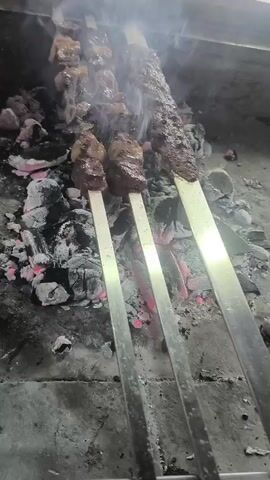 The video shows three metal skewers with pieces of meat being cooked over hot coals. The meat appears to be beef, cut into cubes. Smoke rises from the meat and coals, indicating it is actively cooking. The coals are glowing red and covered in ash. The skewers are positioned over the coals, with the meat about to be cooked. The background is indistinct and appears to be a rough, concrete surface. The scene suggests an outdoor cooking environment, possibly a grill or barbecue. The video is short, focusing on the cooking process.