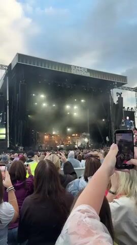 A video clip shows a large outdoor concert stage with the words SUMMER SESSIONS displayed prominently above it. The stage is lit with bright lights, and there is a considerable amount of stage fog. A band is visible performing on stage, with instruments and equipment set up. In the foreground, a crowd of concertgoers is visible, many of them with their arms raised in the air. Some individuals are holding up cell phones, presumably to record or take pictures of the performance. The sky above is cloudy. The video appears to be recorded from the perspective of someone within the crowd.