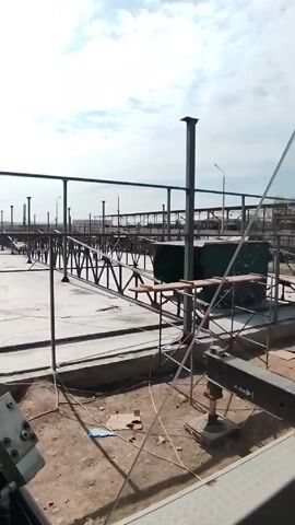 Here's a description of the video:

The video shows a construction site from the perspective of a crane operator. The crane's boom is extending upwards and its cable is visible. Below, a large concrete pad forms the base of the site. Numerous steel support structures, including pillars and crossbeams, are being assembled. In the background, a power line tower is visible against a partly cloudy sky. Workers are present on the construction site, moving around the framework. The ground around the concrete pad is dirt.